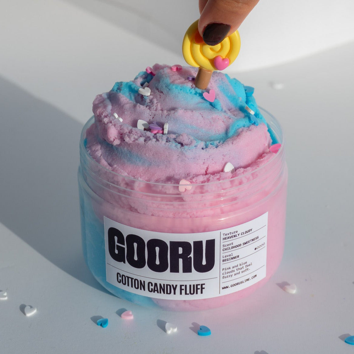 A hand positioning a yellow lollipop charm on top of the Cotton Candy Fluff slime, highlighting the playful decorations and soft swirls.