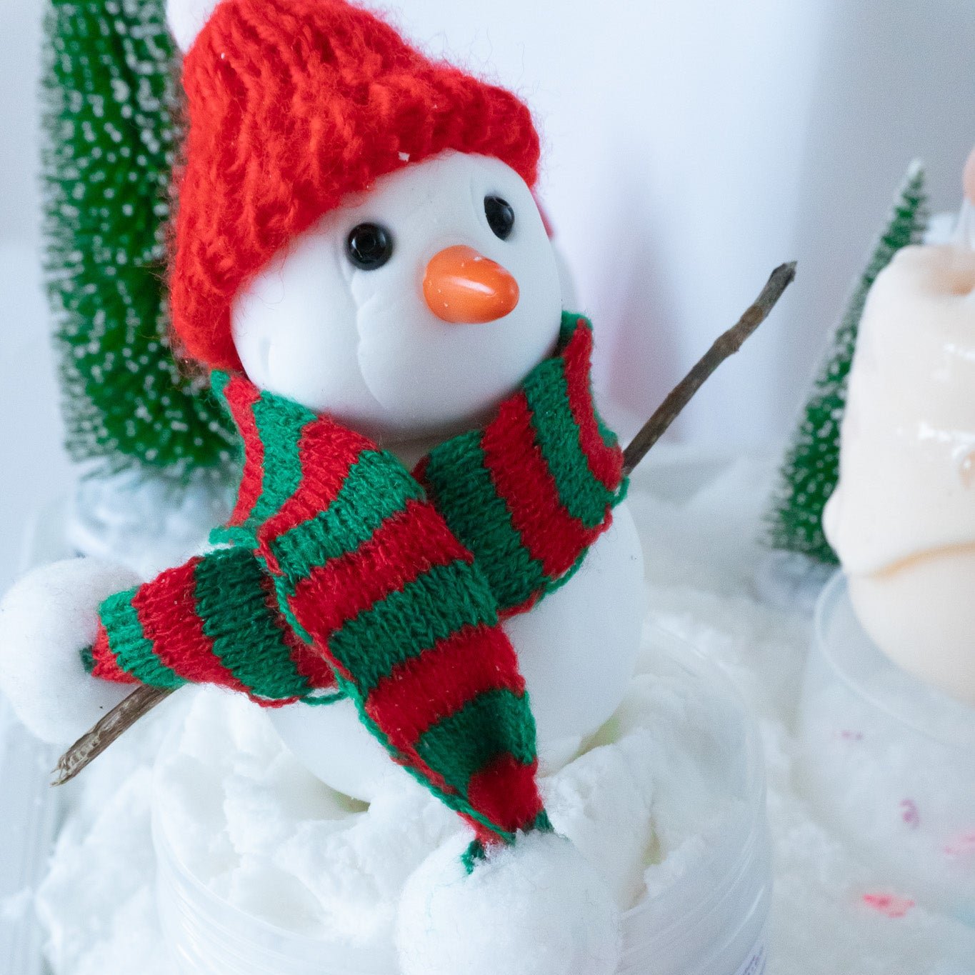 A close-up view of the Build-A-Snowman slime featuring a soft snowman with a red knitted hat and striped green-and-red scarf sitting on fluffy, snow-like slime. Small Christmas trees in the background enhance the festive vibe.