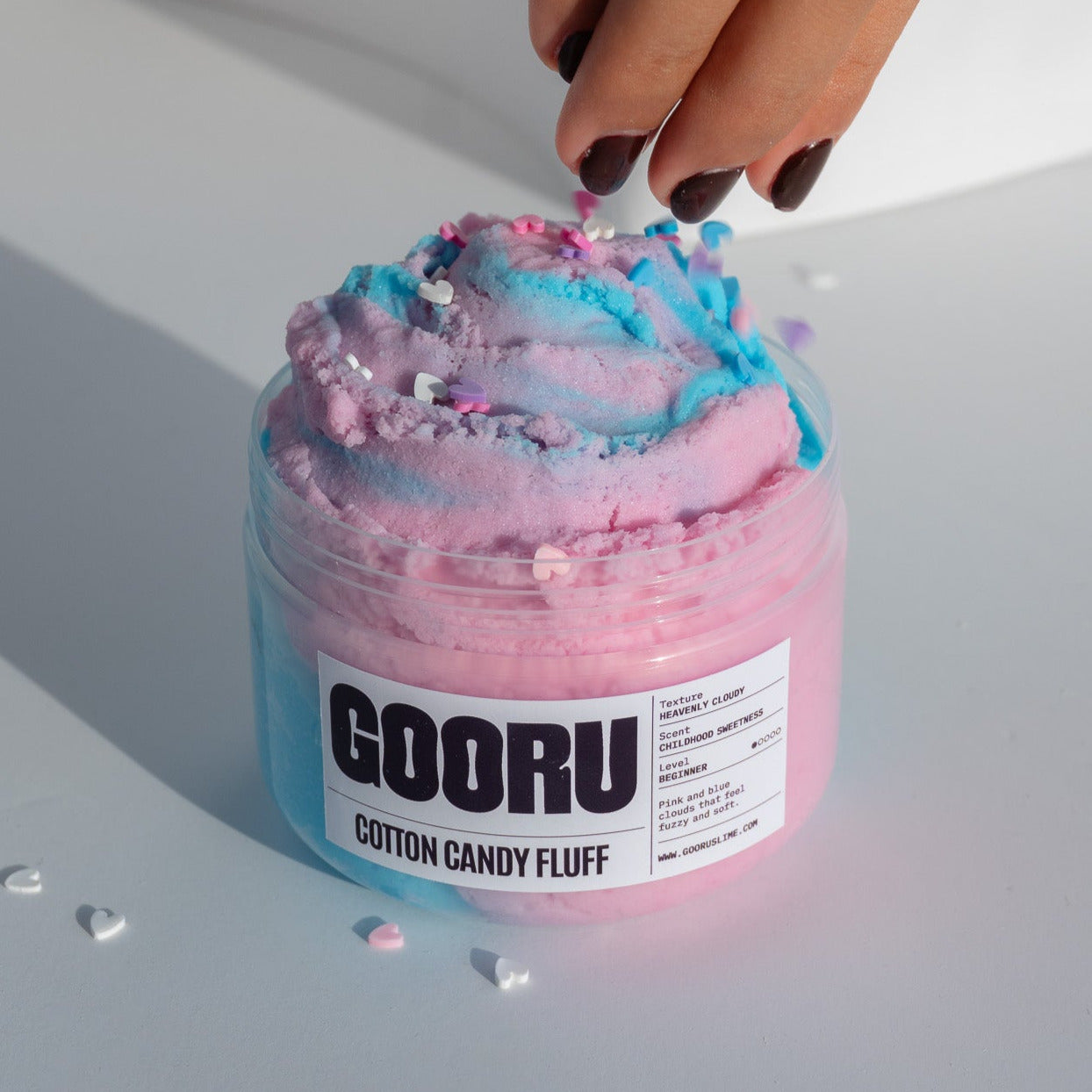  A hand sprinkling colorful heart confetti onto the Cotton Candy Fluff slime, showcasing its cotton candy-inspired design.