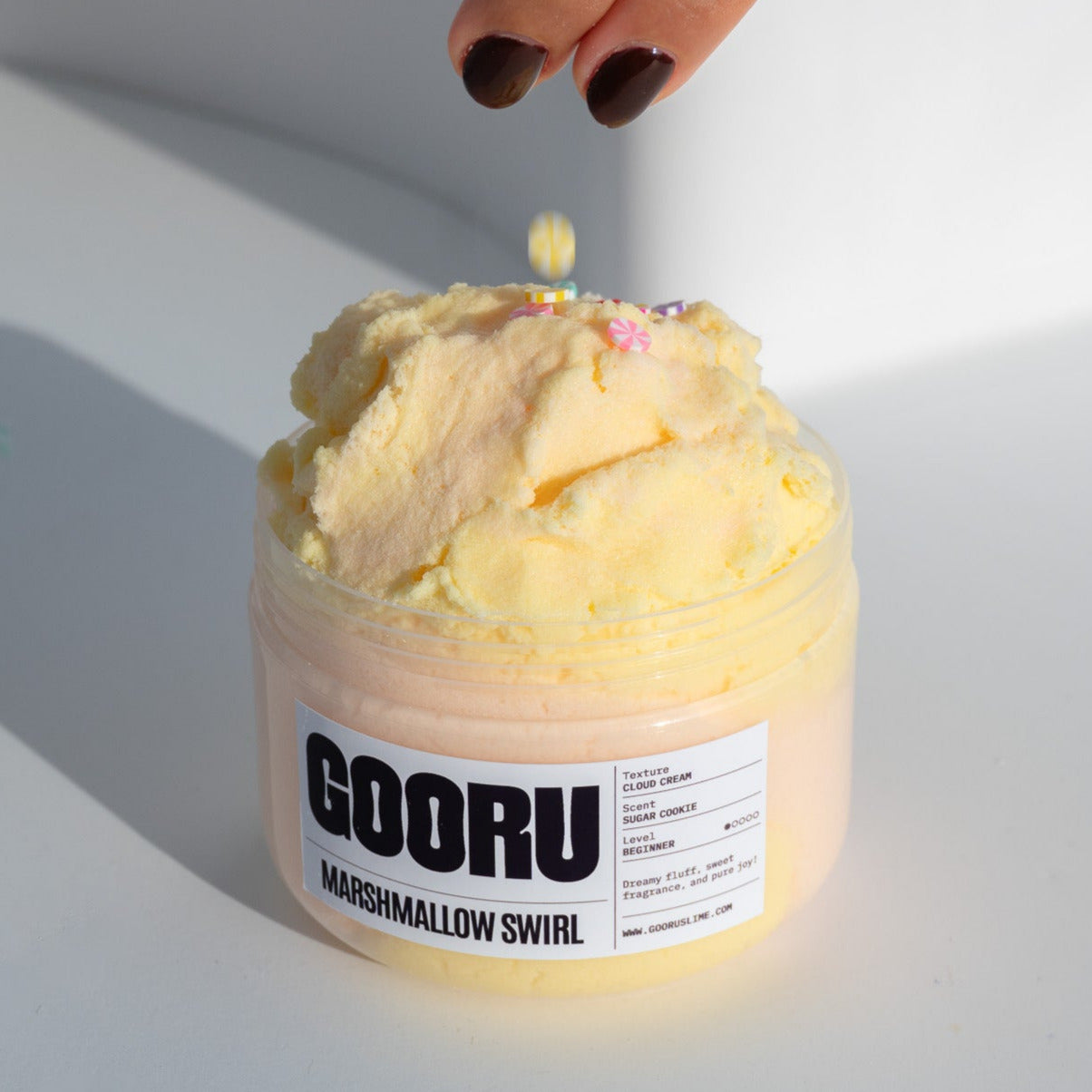 A jar of Marshmallow Swirl slime with a fluffy yellow and pink cloud texture, as a hand sprinkles colorful candy pieces on top.