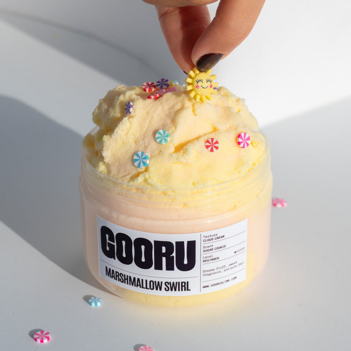 A hand positioning a smiling sun charm onto the Marshmallow Swirl slime, showcasing its creamy, cloud-like texture.