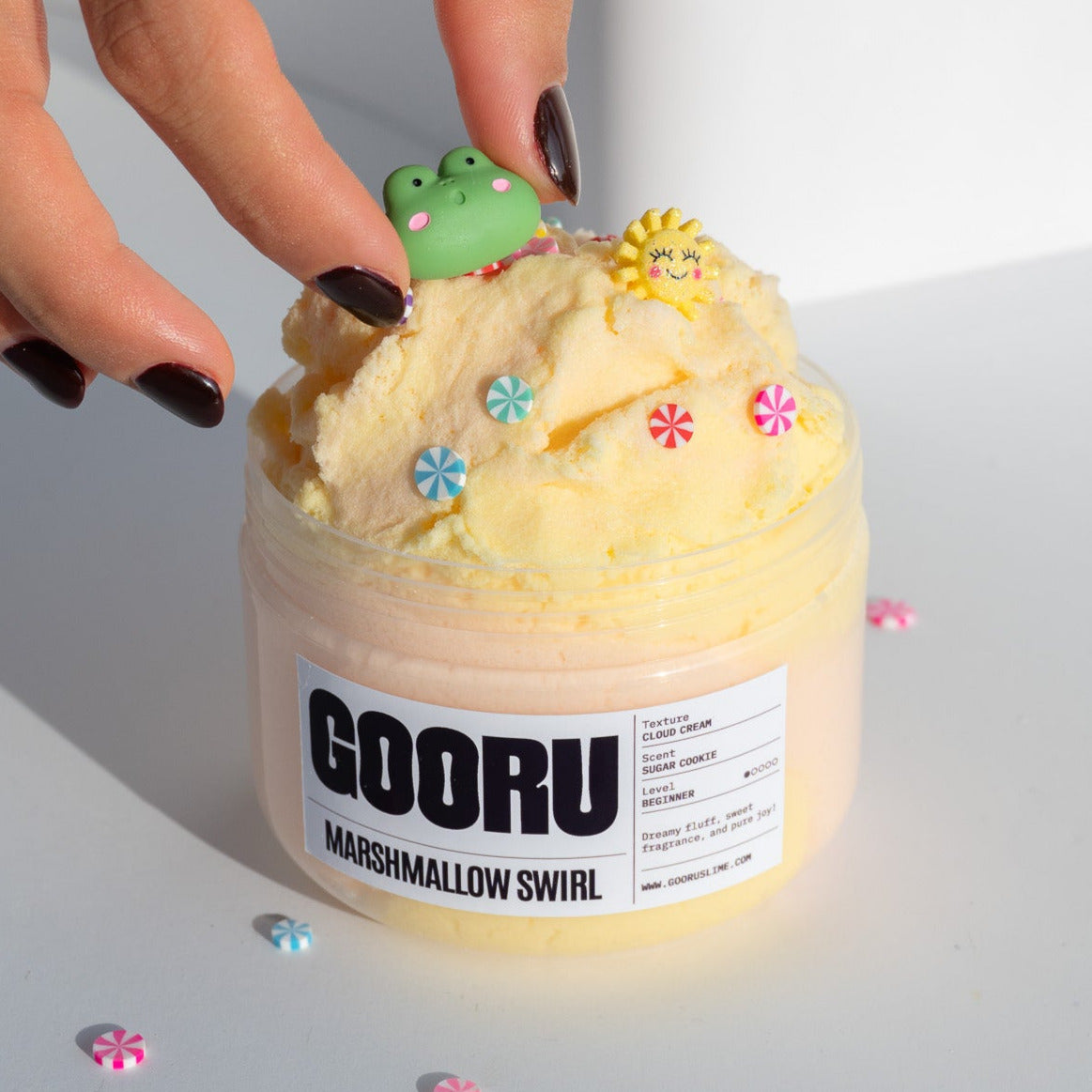 A hand placing a cute green frog charm onto the Marshmallow Swirl slime, surrounded by colorful candy decorations.