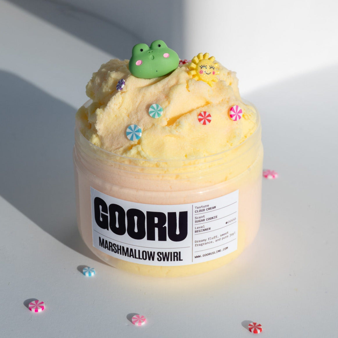 The Marshmallow Swirl slime jar displayed with vibrant yellow and pink tones, adorned with candy and playful charms, including a frog and a sun.