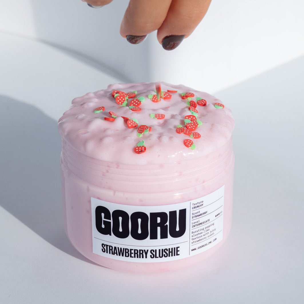 A hand placing a strawberry charm onto the pink, crunchy Strawberry Slushie slime, with bright lighting enhancing the details.
