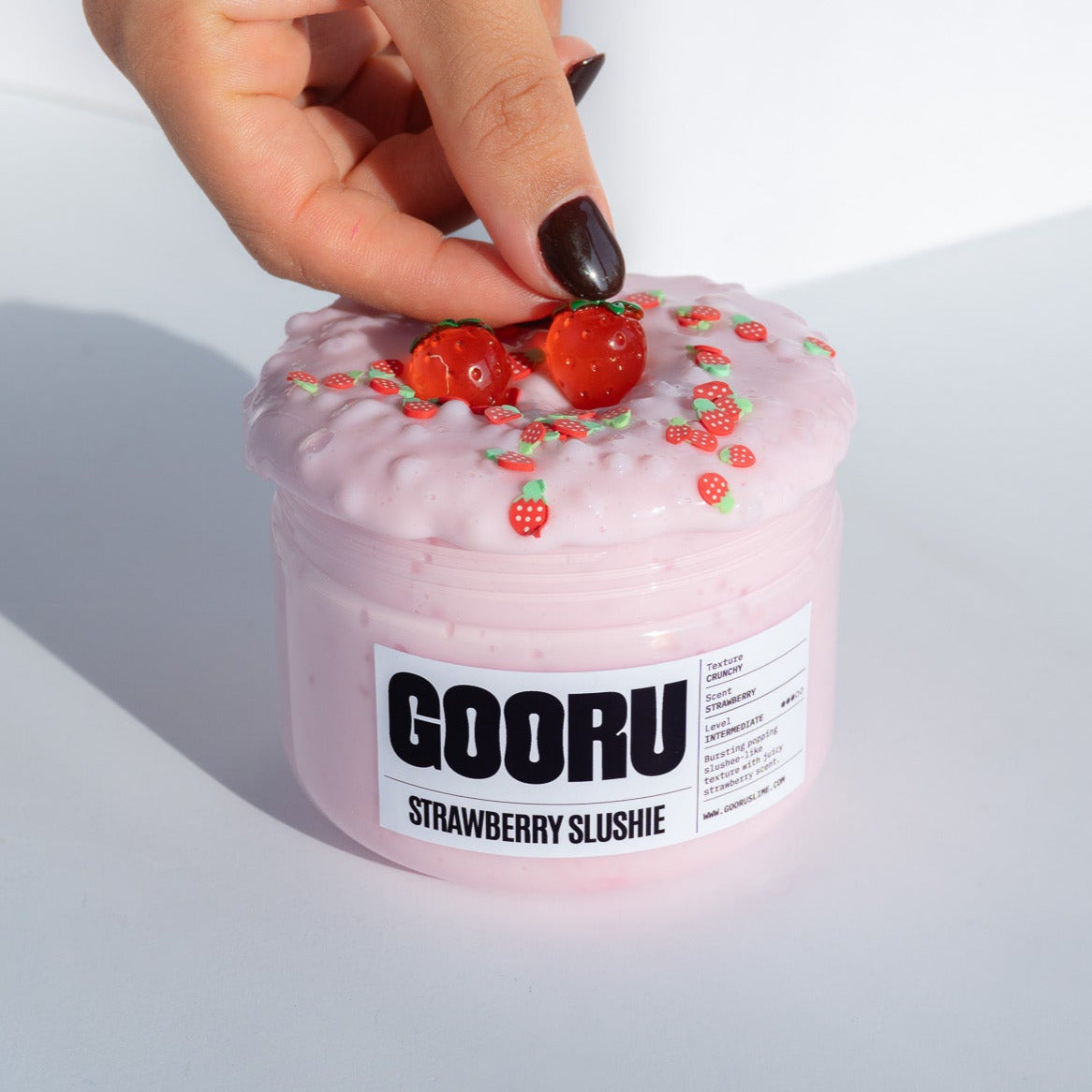 A close-up of a hand adjusting the strawberry charms on the Strawberry Slushie slime jar, showcasing the glossy pink texture.