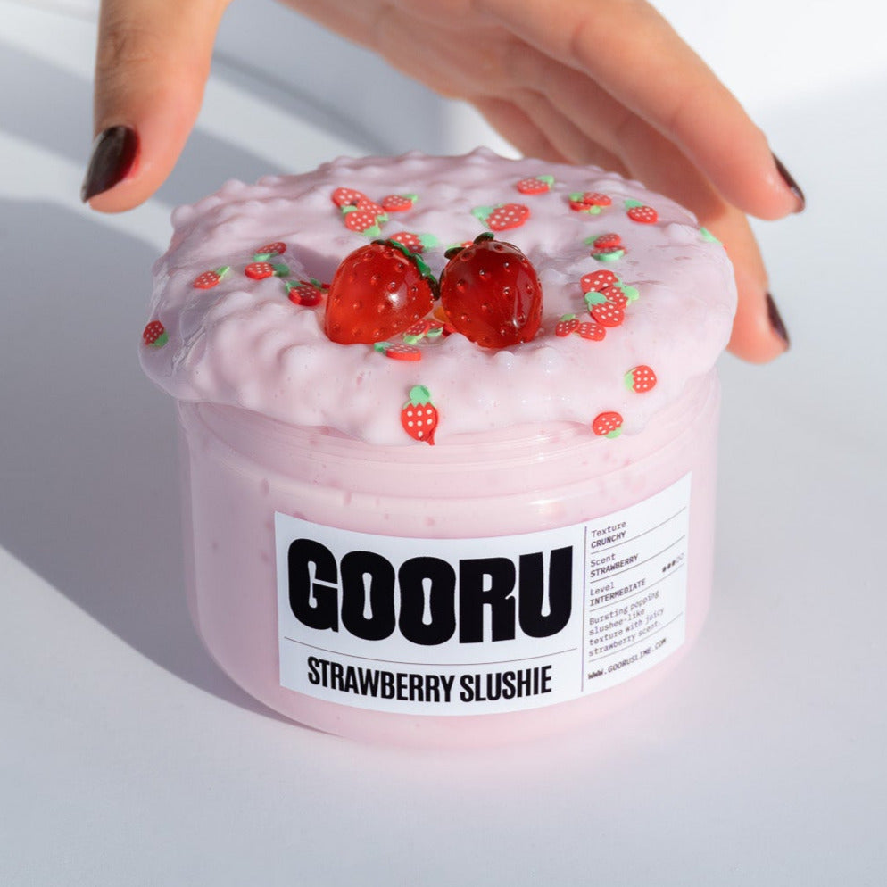 A jar of Strawberry Slushie slime featuring a pink crunchy texture with strawberry charms on top. A hand gently holds the jar.