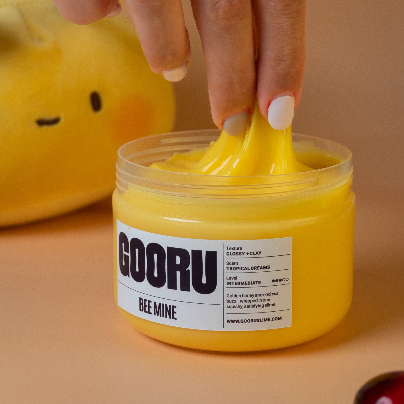 Hand pulling yellow glossy “Bee Mine” slime from the jar with a “GOORU BEE MINE” label.