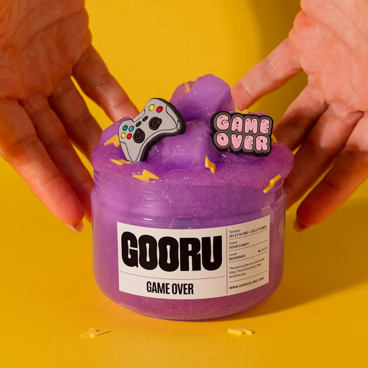 Purple slime container labeled 'GOORU Game Over' held by hands on a yellow background