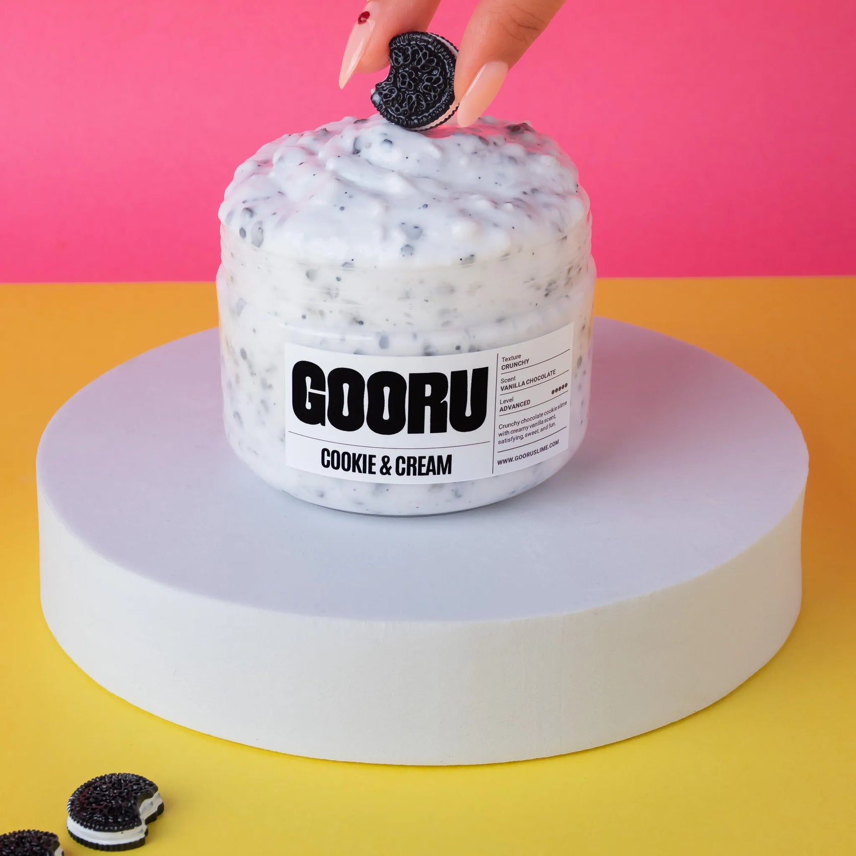 Jar of GOORU Cookie & Cream ice cream with a hand dipping an Oreo cookie into it against a pink and yellow background.
