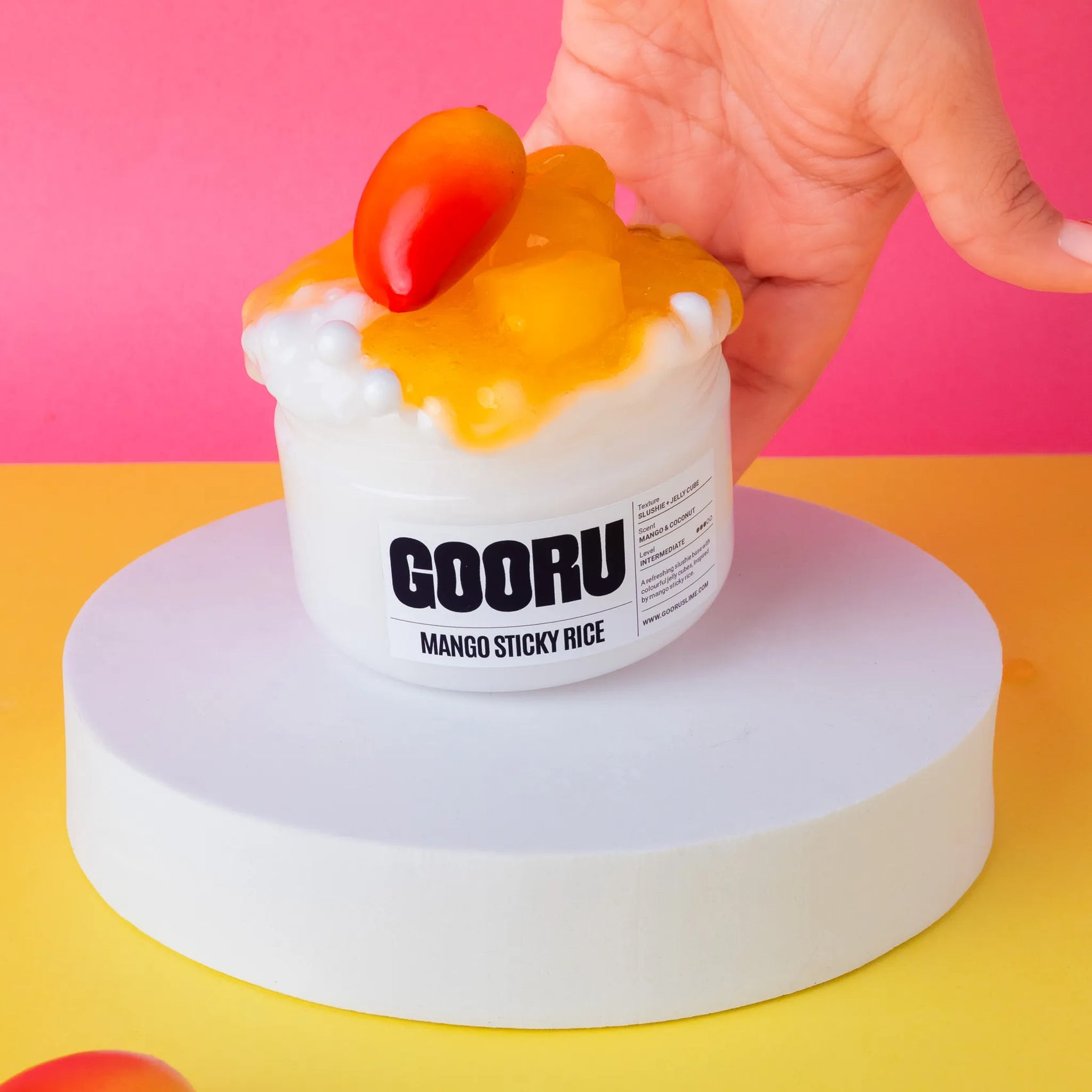 Mango sticky rice slime with mango on top, held by a hand against a pink and yellow background.