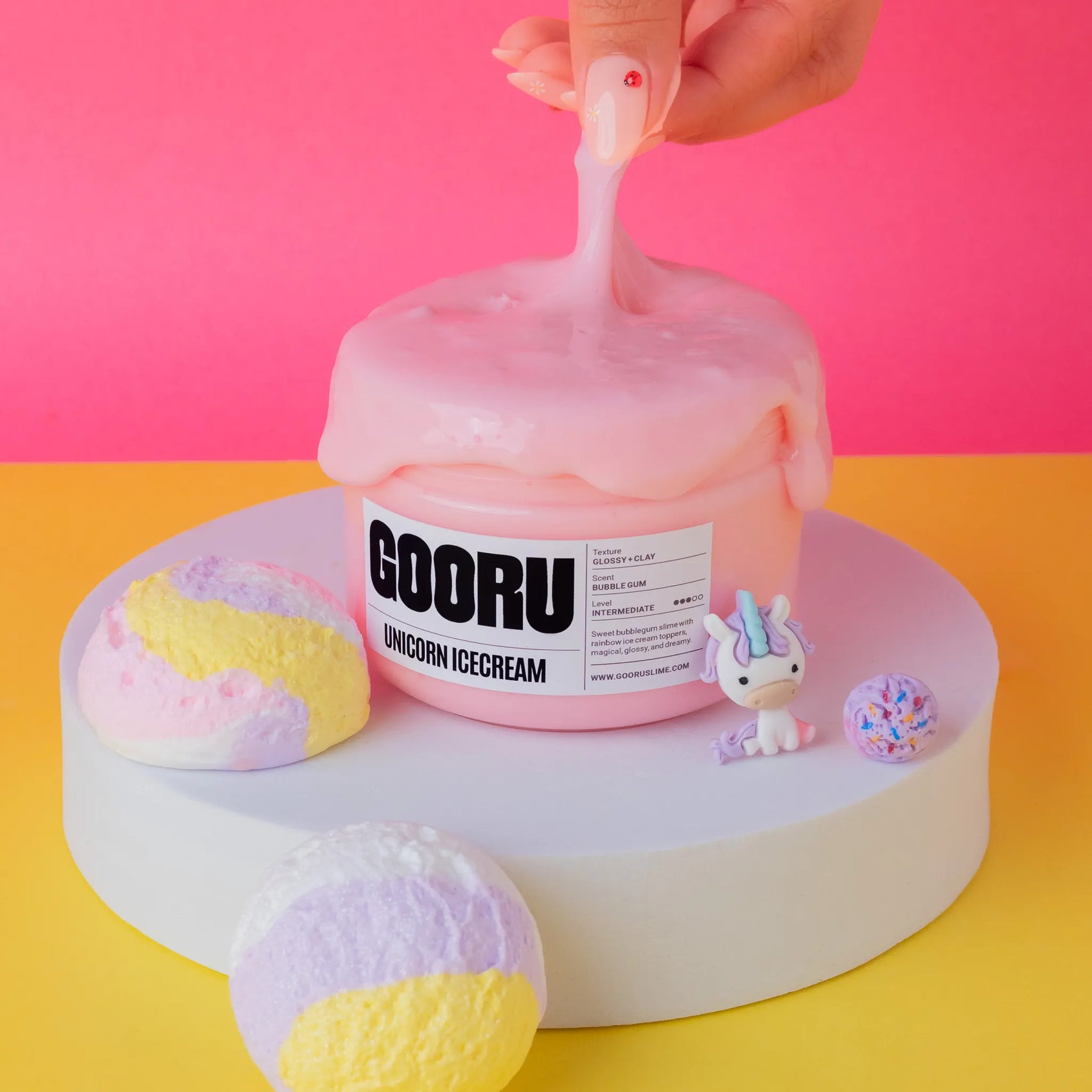 Pink slime container labeled 'GOORU Unicorn Ice Cream' with a hand scooping out pink ice cream against a pink and yellow background.