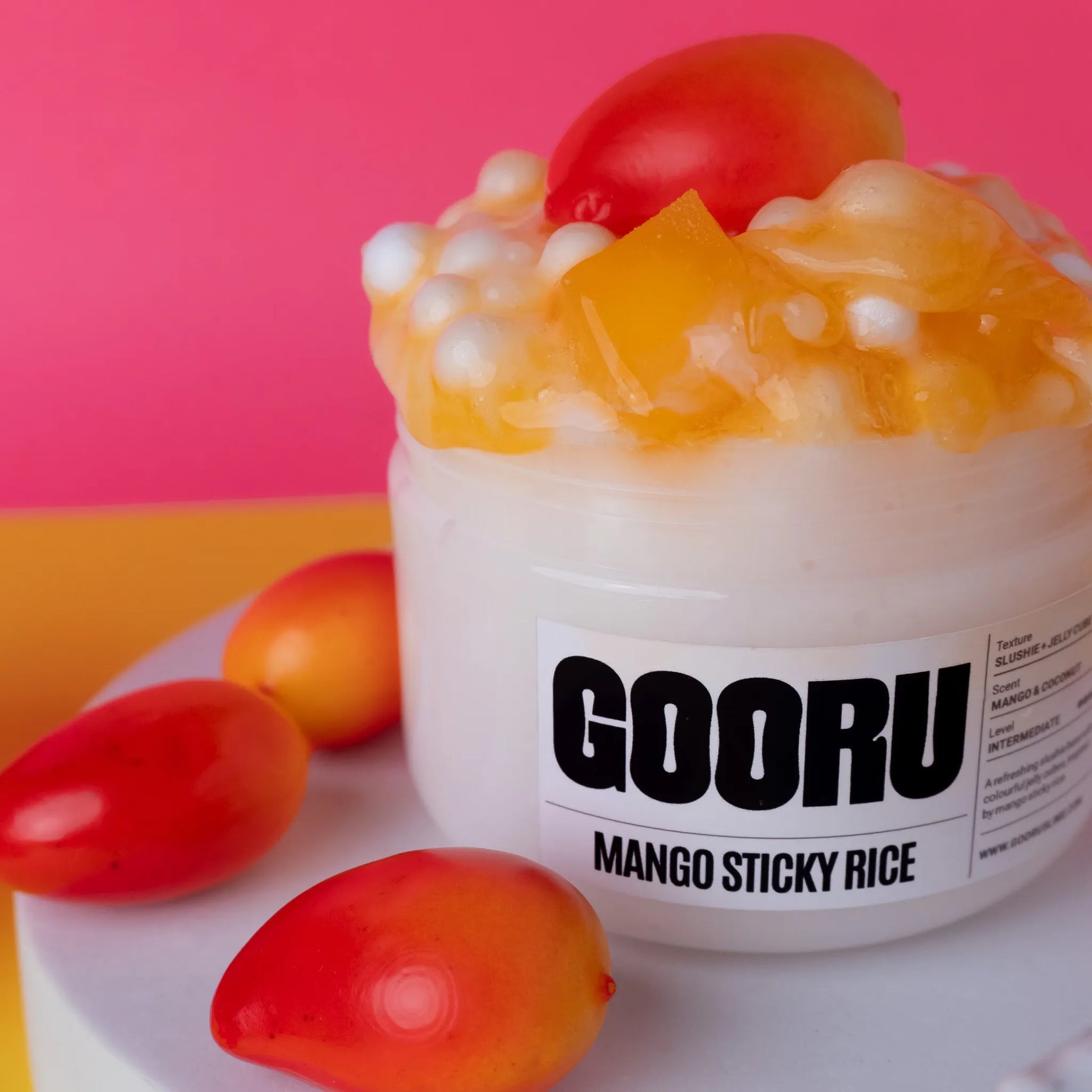 Jar of GOORU Mango Sticky Rice slime with mangoes on a pink background