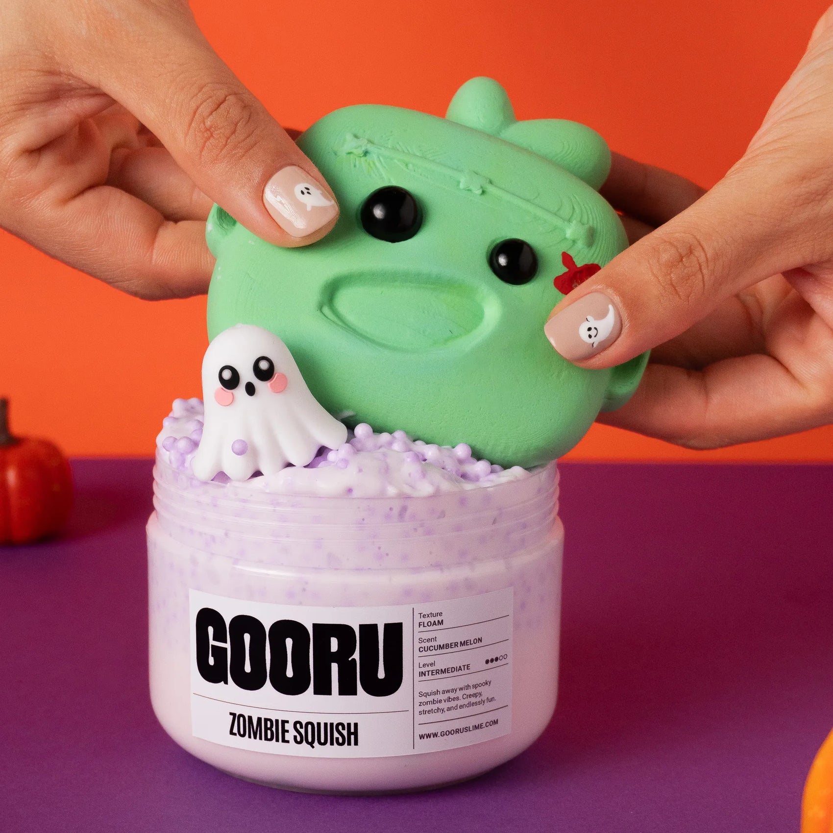 Green zombie squish toy being held above a purple container with a ghost figure, against an orange and purple background.