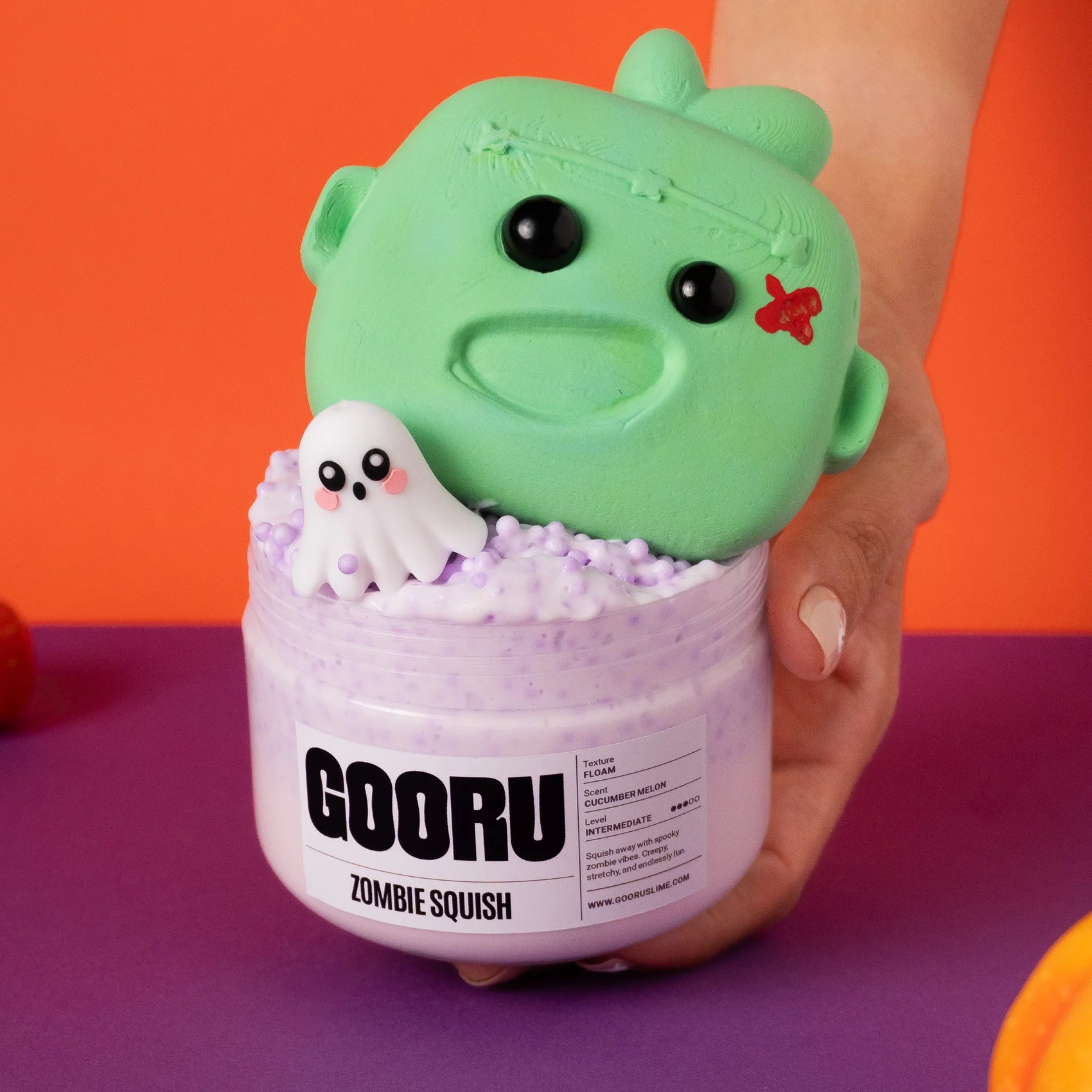 Green zombie squish toy with a ghost inside a container labeled 'GOORU Zombie Squish' on an orange and purple background.