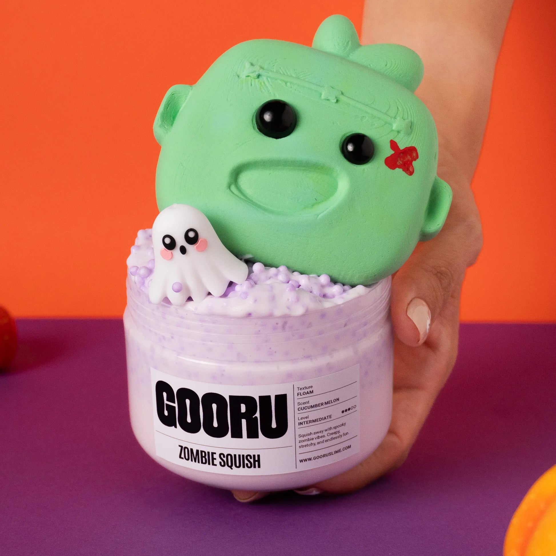 Green zombie squish toy with a ghost inside a container labeled 'GOORU Zombie Squish' on an orange and purple background.