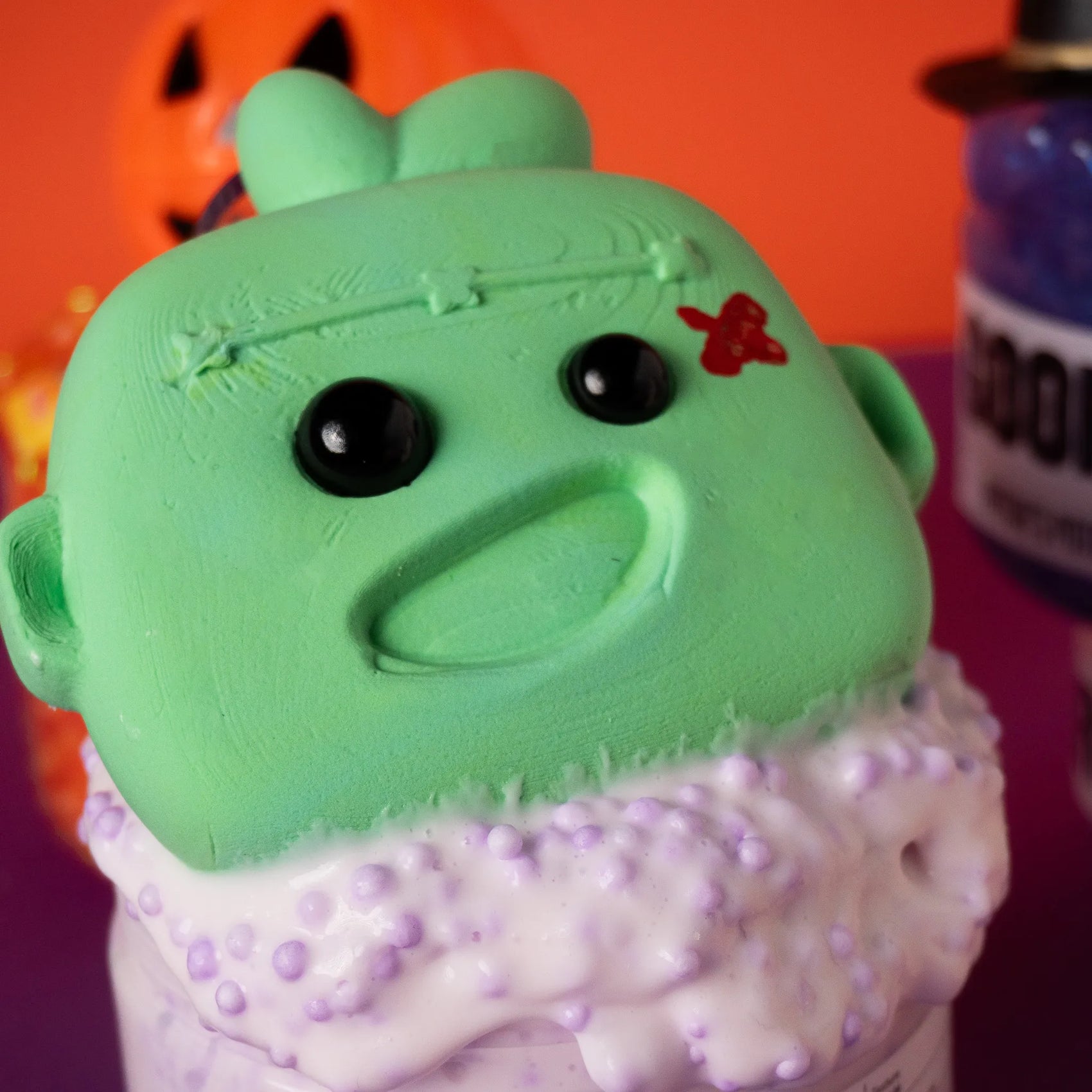 Green and purple toy with black eyes and a red mark on an orange and pink background