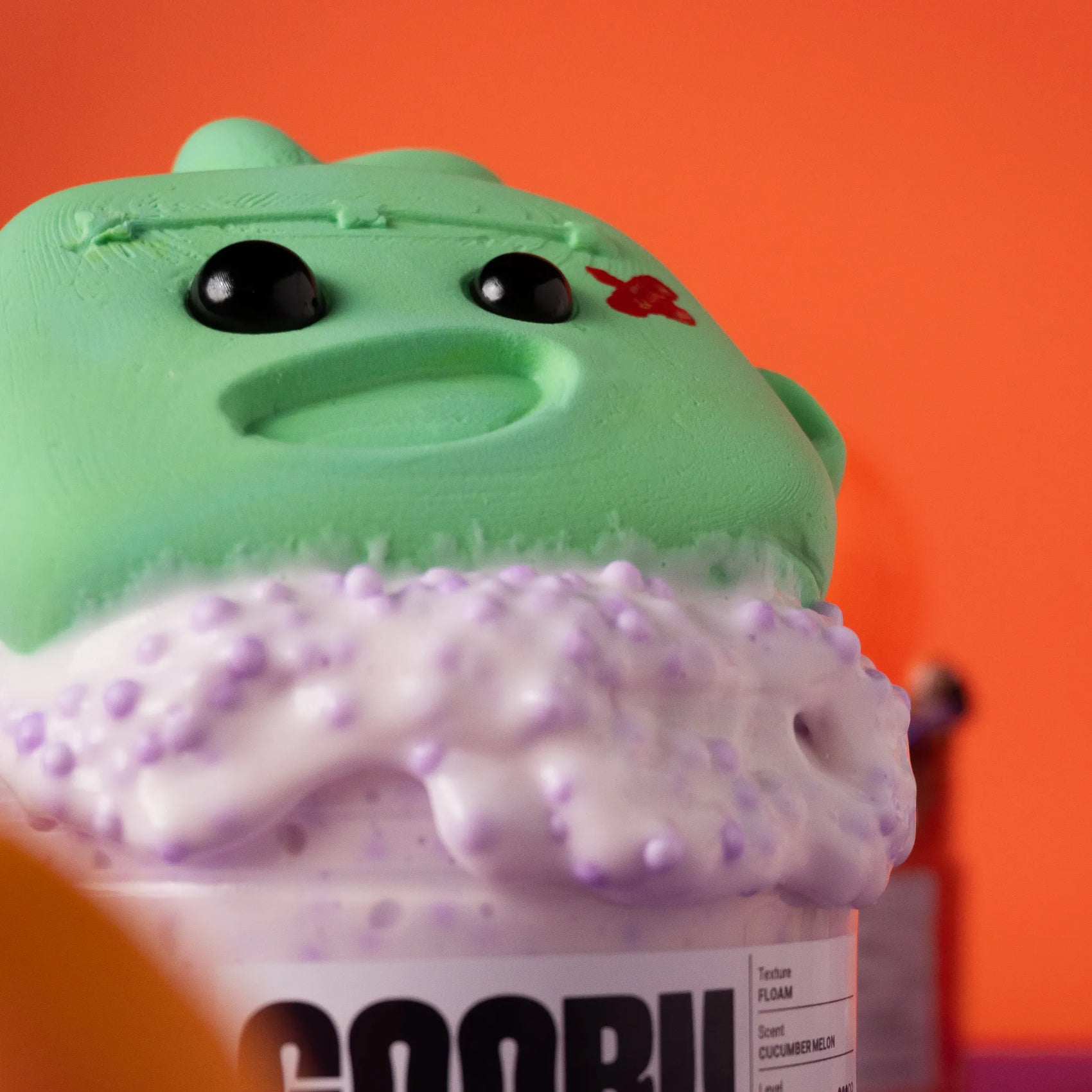 Green and purple textured container with a face on an orange background