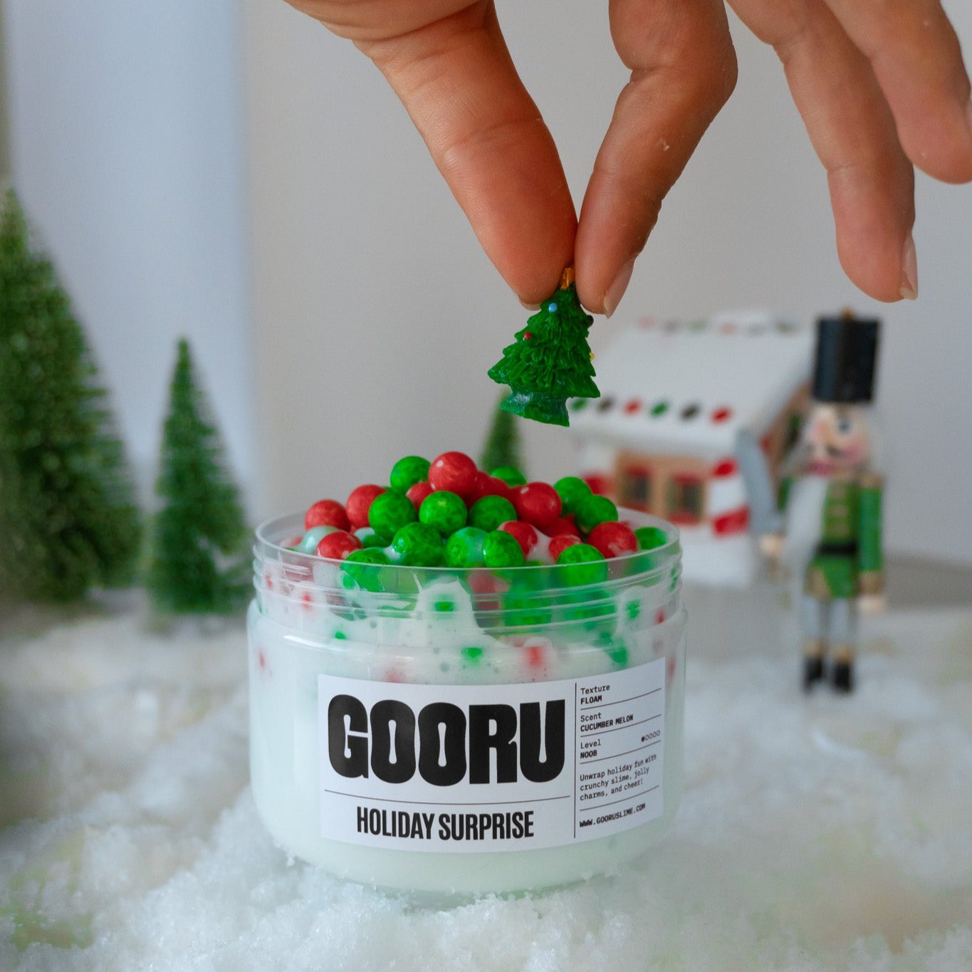 A close-up of Holiday Surprise slime featuring vibrant red and green foam beads with a hand placing a miniature Christmas tree charm on top. The festive background includes snow and a nutcracker figurine.